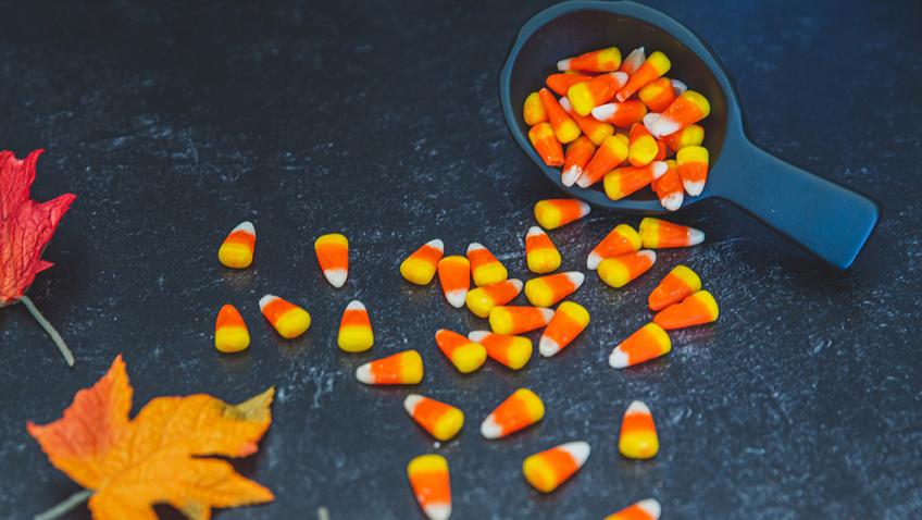 Candy corn spills out of a dark gray measuring cup onto a marble surface next to colorful fall leaves