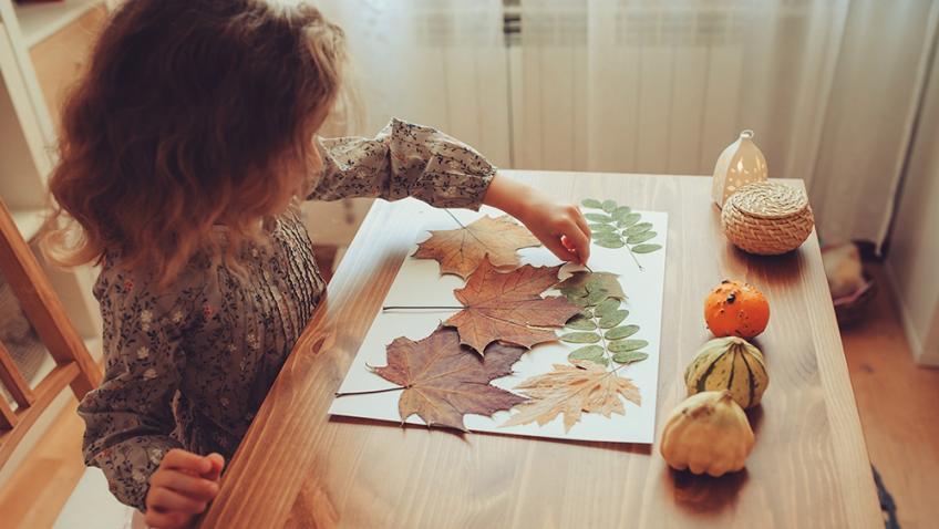 A child sitting at a kitchen table glues leaves to a piece of craft paper