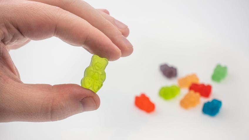 A hand holds a yellow gummy bear between thumb and forefinger in front of a small pile of multicolored gummy bears