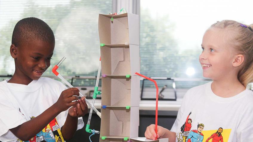Two elementary students work together to connect clear plastic pipes in a circuit around a rectangular tower of cardboard