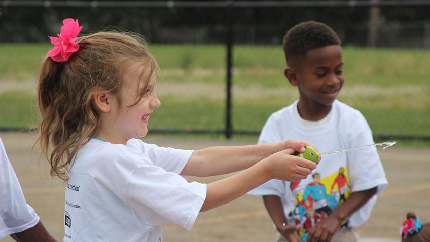 A child stands in an outdoor baseball field and uses a toy fish to squirt a stream of water at another child
