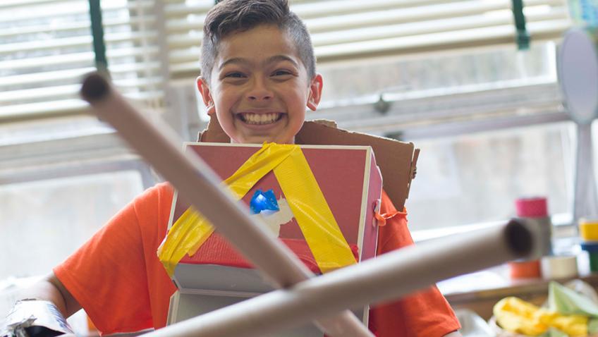 A grinning elementary student crosses two long cardboard tubes in front of themself