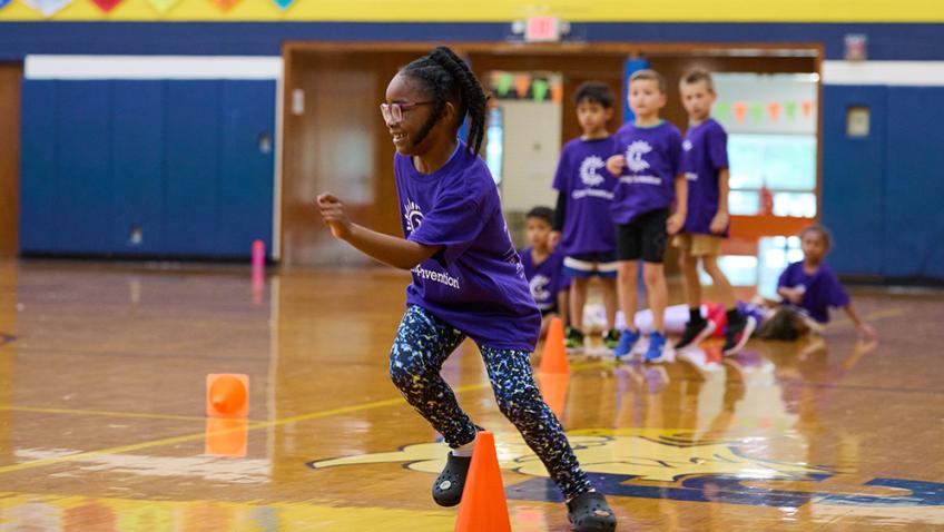 An elementary student wearing a purple Camp Invention shirt sprints around a cone in a school gymnasium