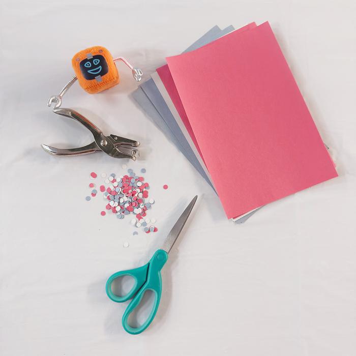 Colorful sheets of craft paper in a stack, a metal hole punch, scissors and a small pile of colorful paper circles