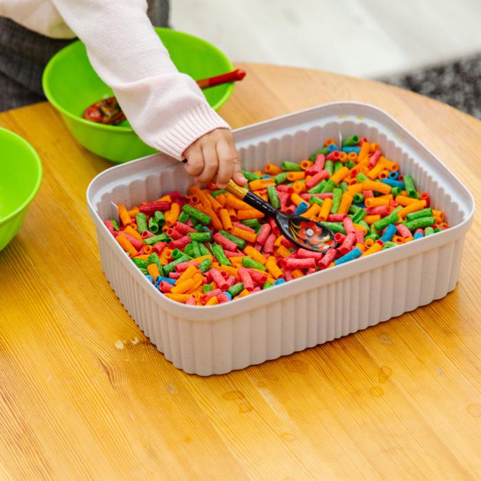 A small child dips a wooden spoon into a large white plastic bin full of multicolored dry pasta