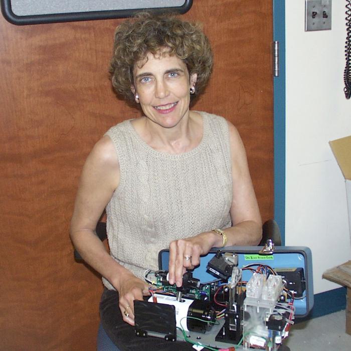 Frances Ligler sits in a chair and holds in her lap an opened biosensor that has the internal components exposed