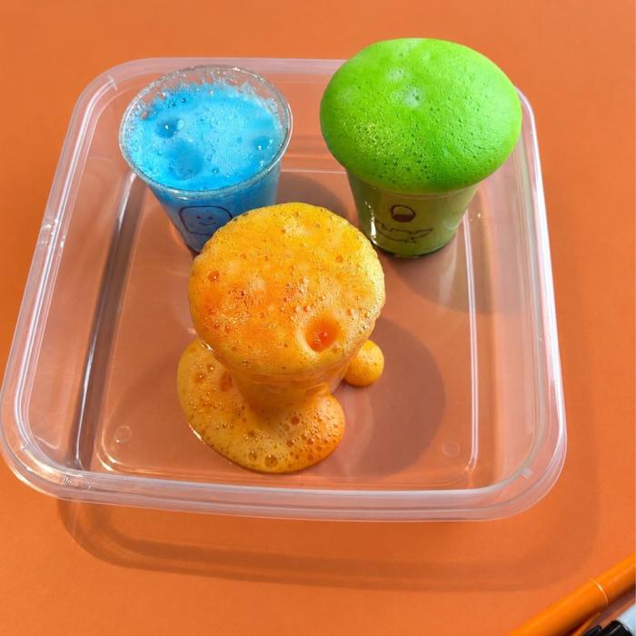 Three cups overflowing with blue, green and orange foam sitting inside a clear plastic container