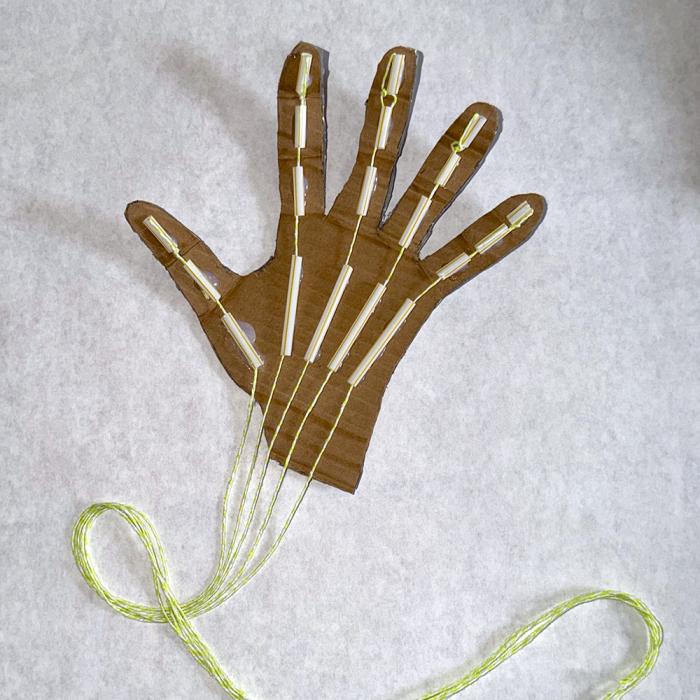 A hand cut out of cardboard with pieces of a straw attached to each finger and string threaded through them