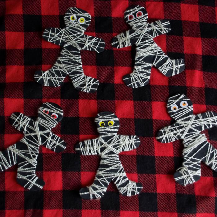Dark cardboard cutouts wrapped with white yarn to look like mummies sit on top of a red plaid tablecloth