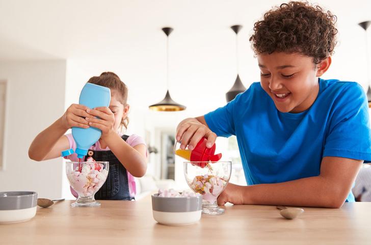 Two children add sprinkles and strawberry syrup to cups of ice cream topped with marshmallows