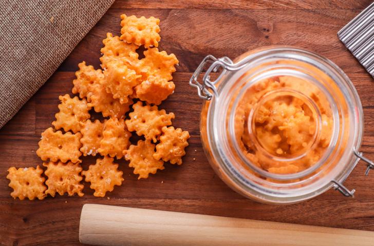 Homemade cheddar crackers spread on a wooden table next to a rolling pin and a glass jar filled with more crackers