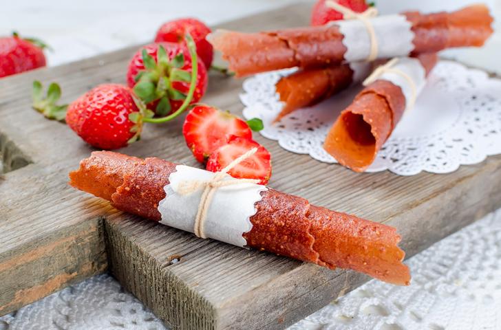 Homemade fruit strips rolled into tubes and tied with parchment paper and twine sit on a cutting board with strawberries