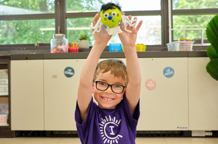 An elementary student wearing a purple Camp Invention shirt holds up a small, green, round robot