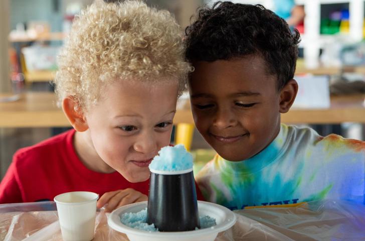 Two children lean their heads together and look down at bright blue foam spilling out of a black paper volcano