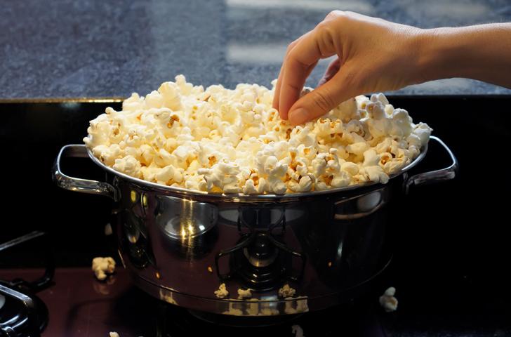 A hand reaches out to grab a piece of popcorn out of a pot filled with popcorn sitting on a stovetop