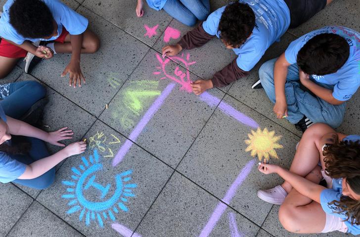 A group of children sits on a cement patio and draws a colorful Camp Invention themed border with sidewalk chalk