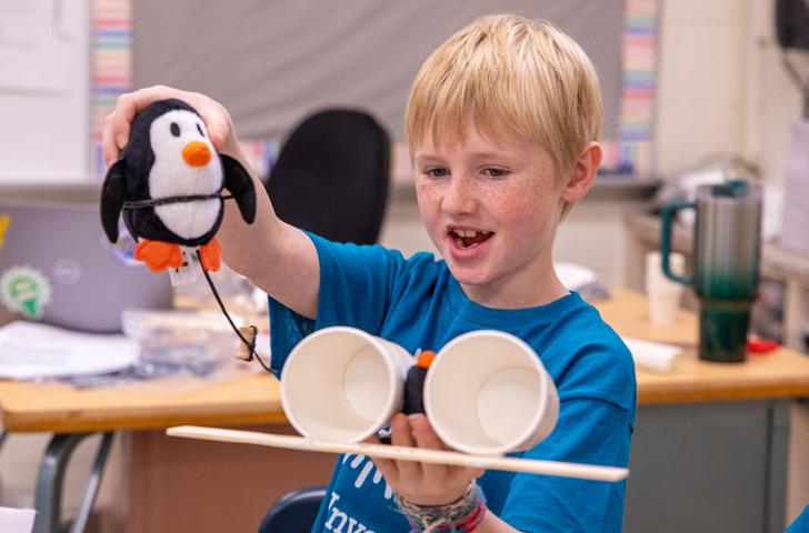 A Camp Invention camper holds up a penguin plushie connected by a string to the camper’s invention prototype