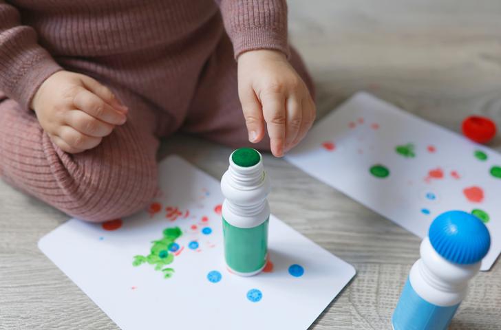 A small child reaches for an opened green dot marker sitting on top of a paper covered in colorful dots