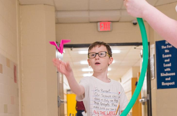 An elementary student tosses a paper airplane through a hula hoop that another student is holding up 