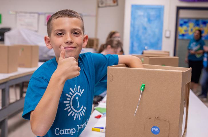 A Camp Invention camper pauses to give a thumbs up before continuing to work on their invention prototype