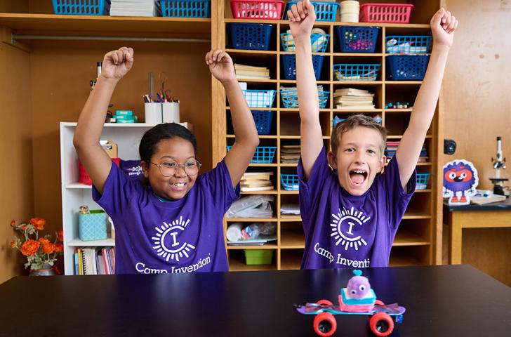 Two elementary students cheer loudly as a small mobile robot rolls across a desk