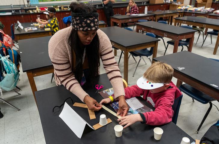 An educator helps a student measure a piece of tape to attach paper cups and cardboard to create an invention prototype