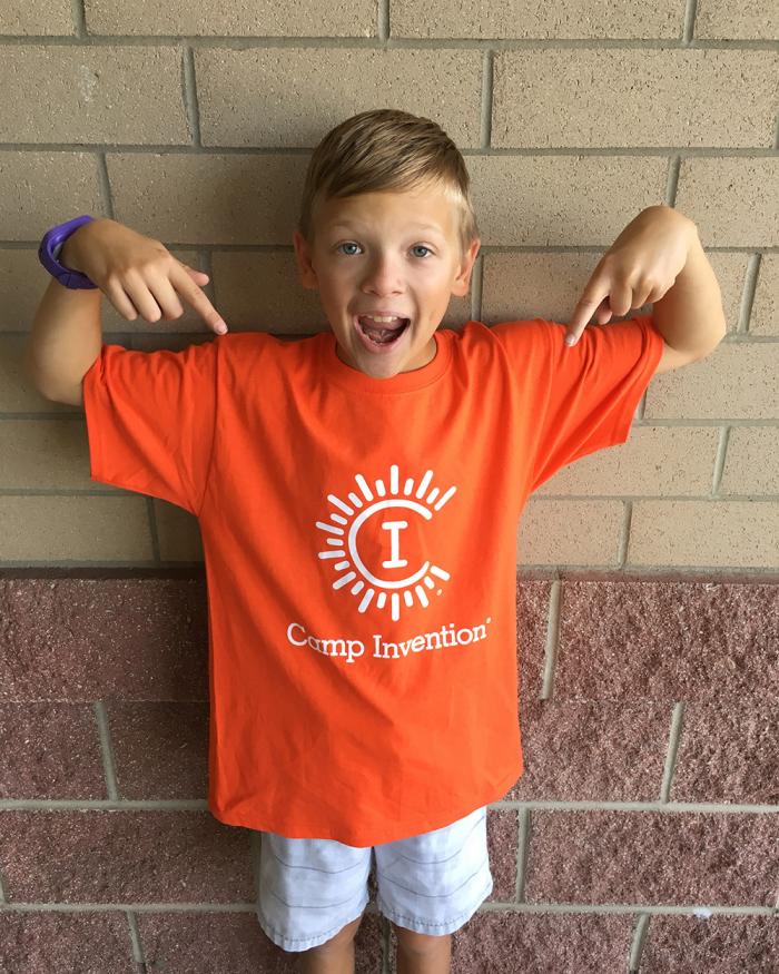 Aydin Wilhelm as an excited child pointing at the orange Camp Invention shirt he is wearing