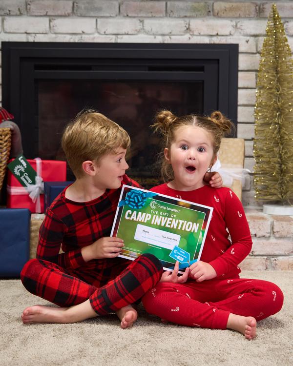 Two siblings wearing red pajamas sit on the floor and excitedly hold up a certificate for a Camp Invention registration