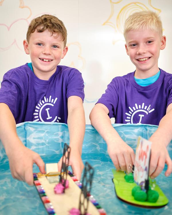 Two Camp Invention campers smile as they place the miniature billboard boats they created into a small pool of water