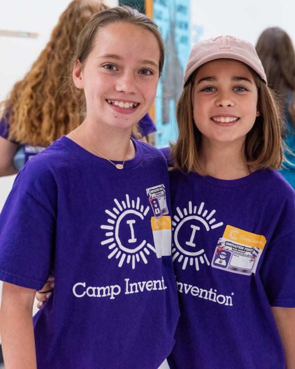 Two siblings wearing matching Camp Invention shirts smile and put their arms around each other