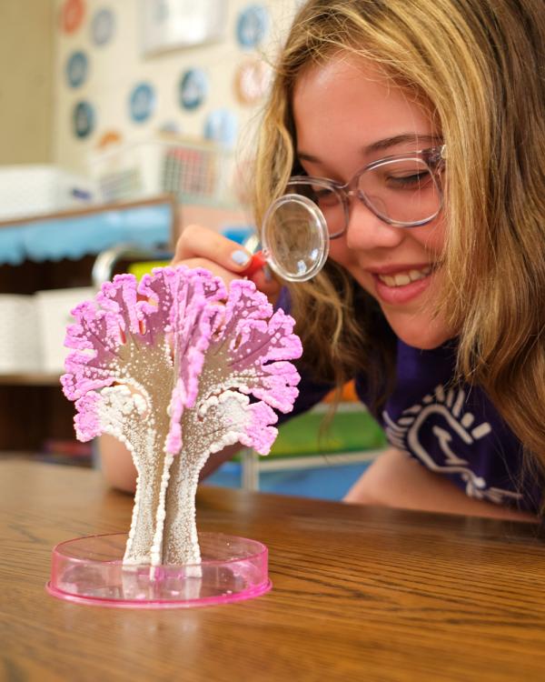 A Camp Invention camper leans forward and uses a small plastic magnifying glass to look closely at a pink crystal tree