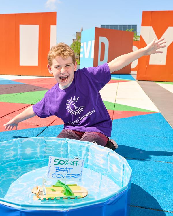 A Camp Invention camper grins and spreads their arms wide as they sit behind their miniature billboard boat