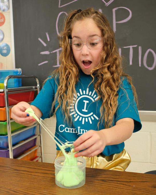 An excited elementary student wearing a blue Camp Invention shirt pulls light green slime out of a clear plastic container