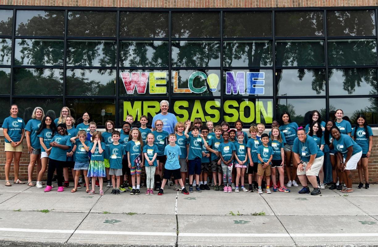 Steve Sasson and a group of elementary students stand in front of windows painted with the words “Welcome Mr. Sasson”